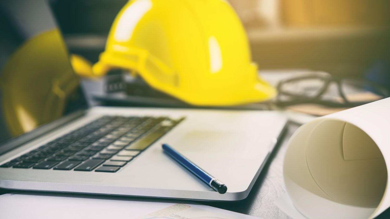 Construction payroll workspace with a laptop, yellow hard hat, blueprints, and pen.