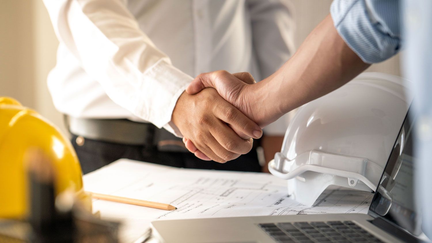 Two construction professionals shaking hands over blueprints with safety helmets and a laptop on the desk.