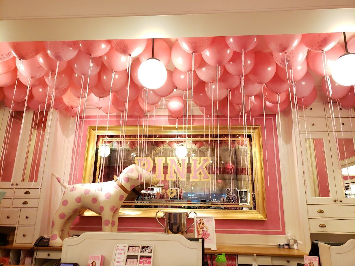 Pink balloon-filled room with a dog statue and a bright neon “PINK” sign on the wall.