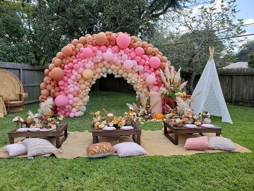 Pink balloon arch over a backyard tea party setup with cushions, low tables, and a teepee on grass