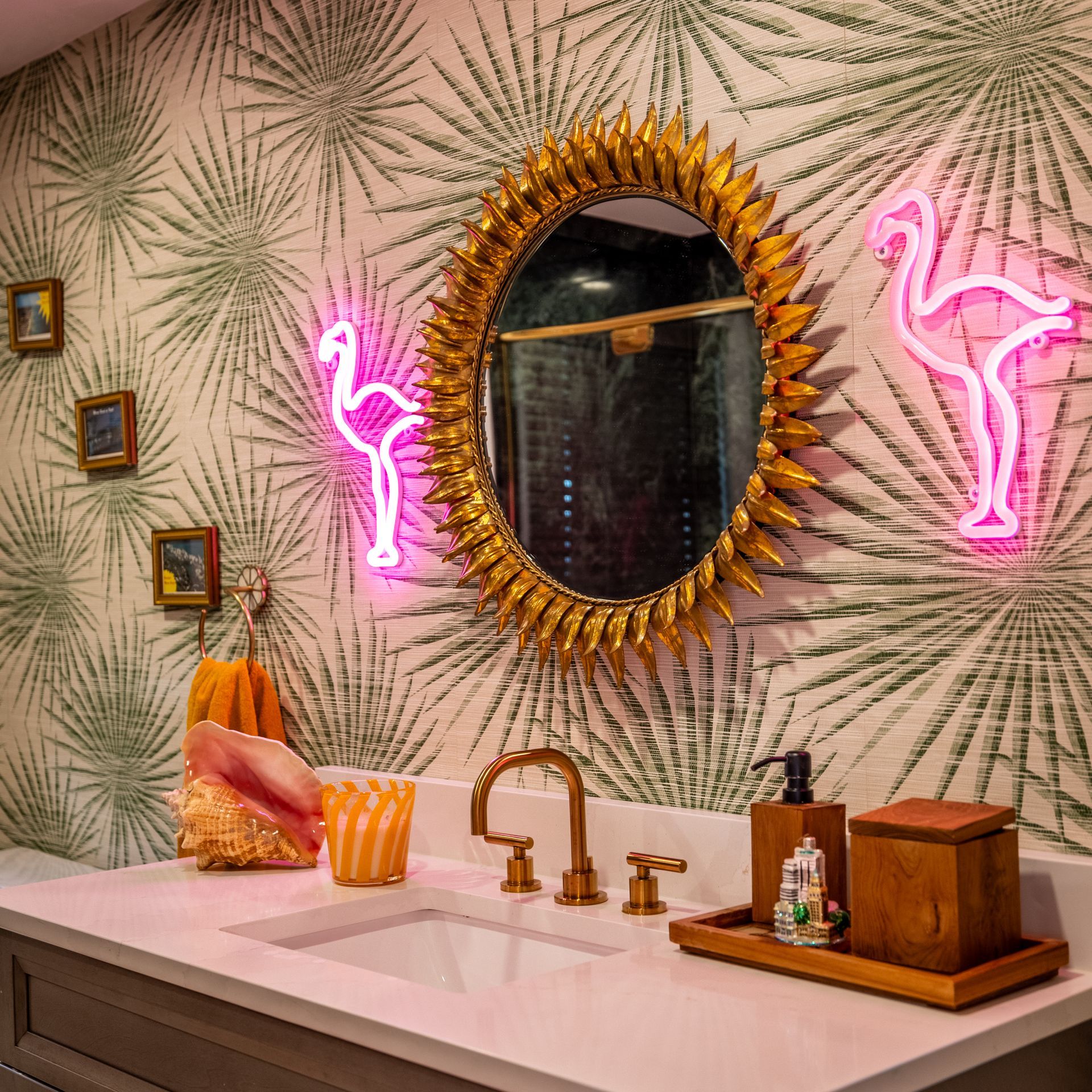 Bathroom with patterned wallpaper, gold mirror, neon flamingo lights, and sink with gold fixtures.