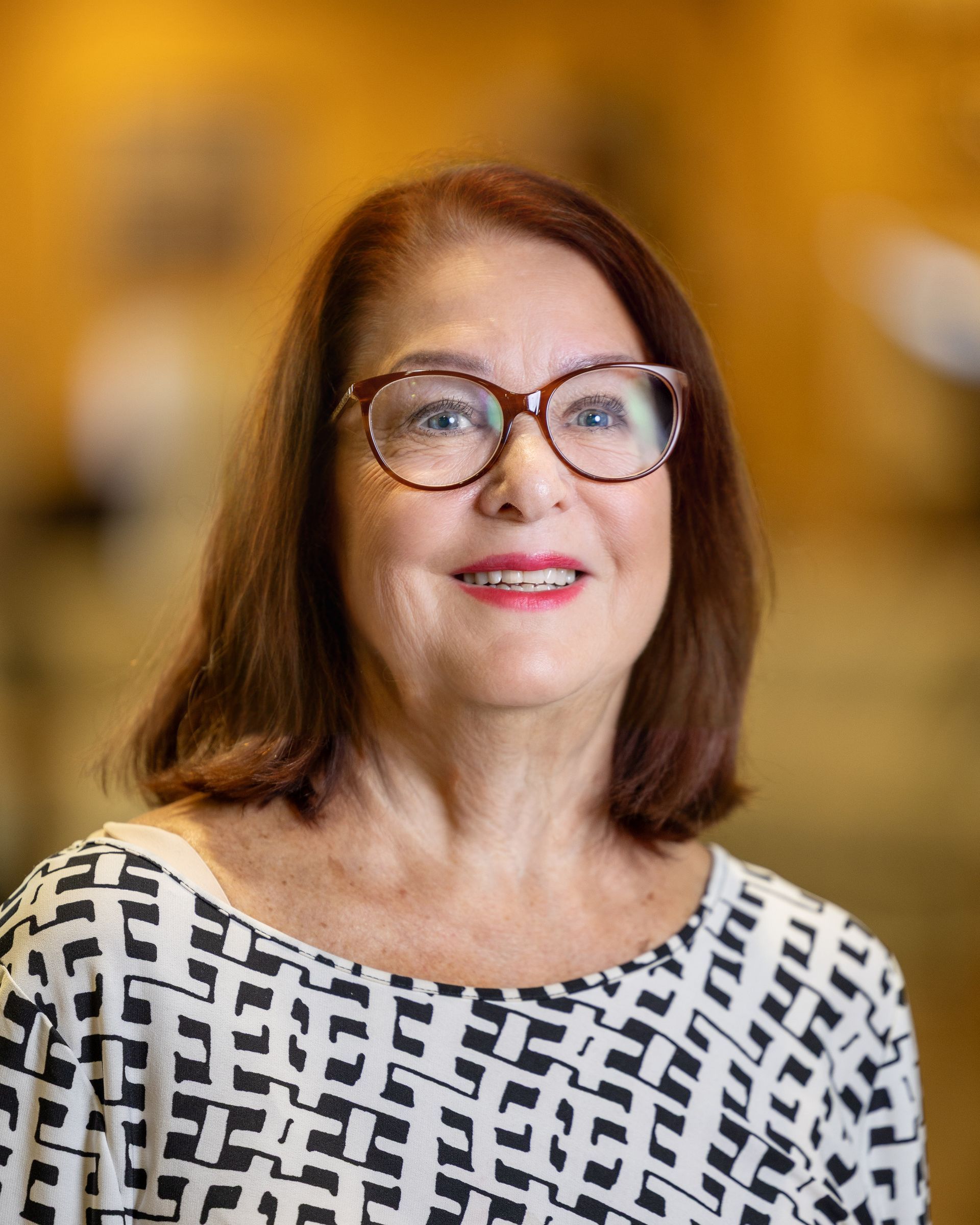 Woman wearing glasses, smiling, with short brown hair, in a black and white patterned top. Blurred indoor background.