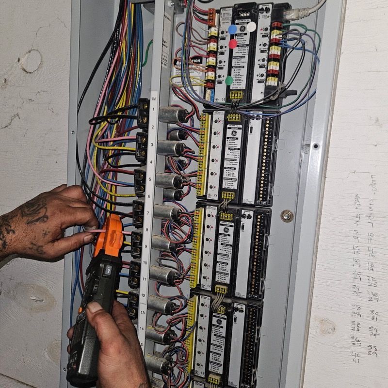 A technician uses a clamp meter to test electrical wires inside a commercial lighting control panel.