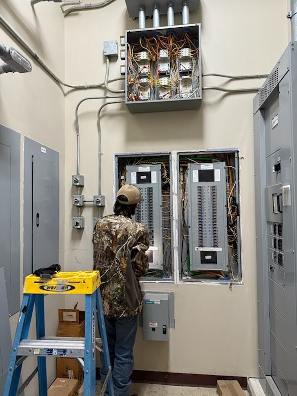 An electrician in a camo jacket works on two open electrical breaker panels with a yellow ladder in the foreground.