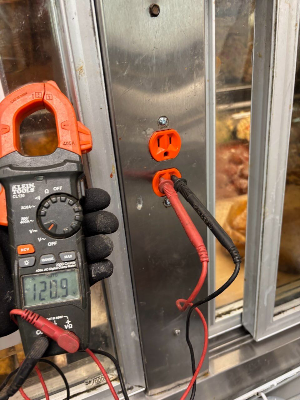 A gloved hand holds a digital multimeter showing 120.9 volts while testing an orange electrical outlet on a metal frame.