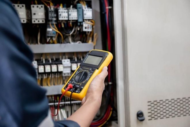 A person in a blue uniform holds a yellow digital multimeter in front of an open electrical control panel.