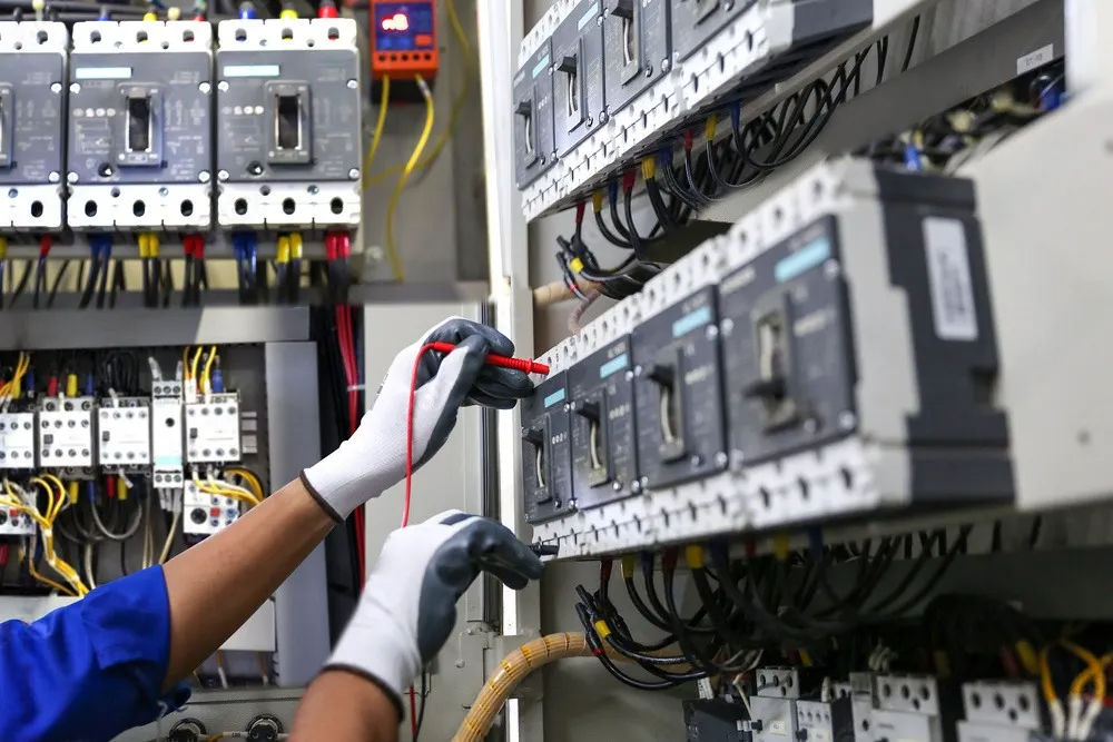 A person in protective gloves uses test probes to inspect electrical components inside an industrial circuit breaker panel.