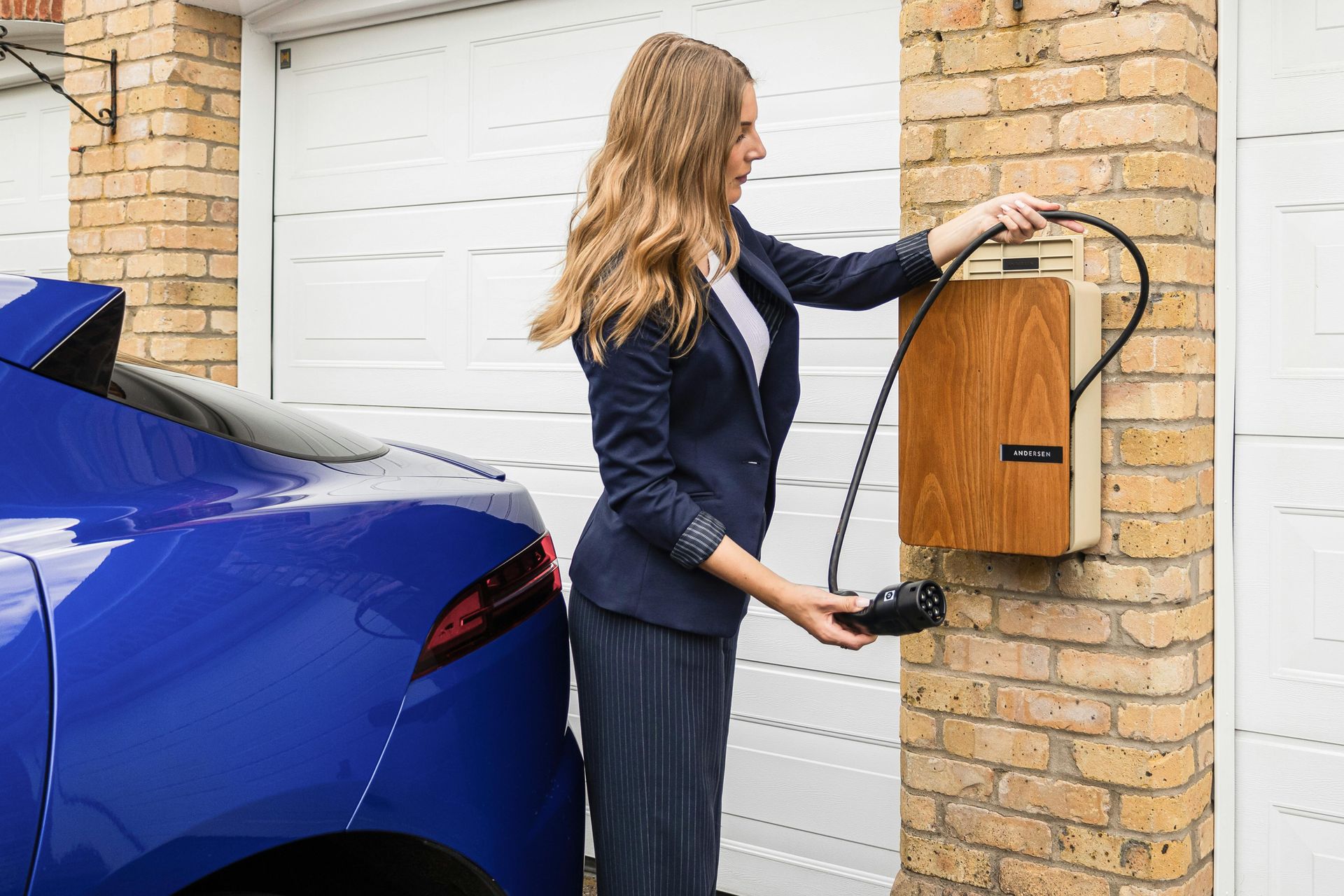 A person in professional attire plugging a charger into a blue electric car parked in front of a brick garage.