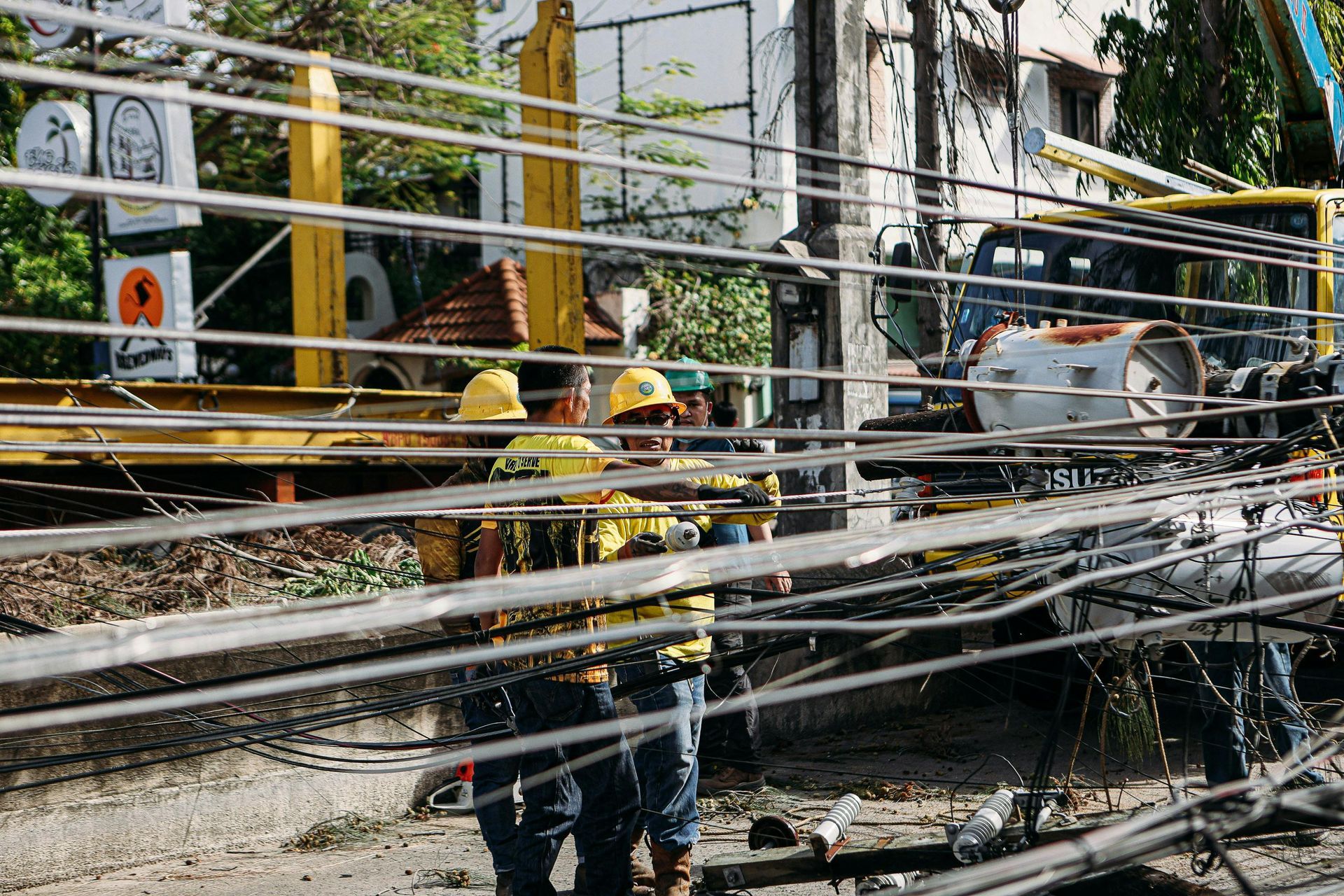 Two utility workers in yellow safety gear and hard hats work on electrical power lines near a utility truck.