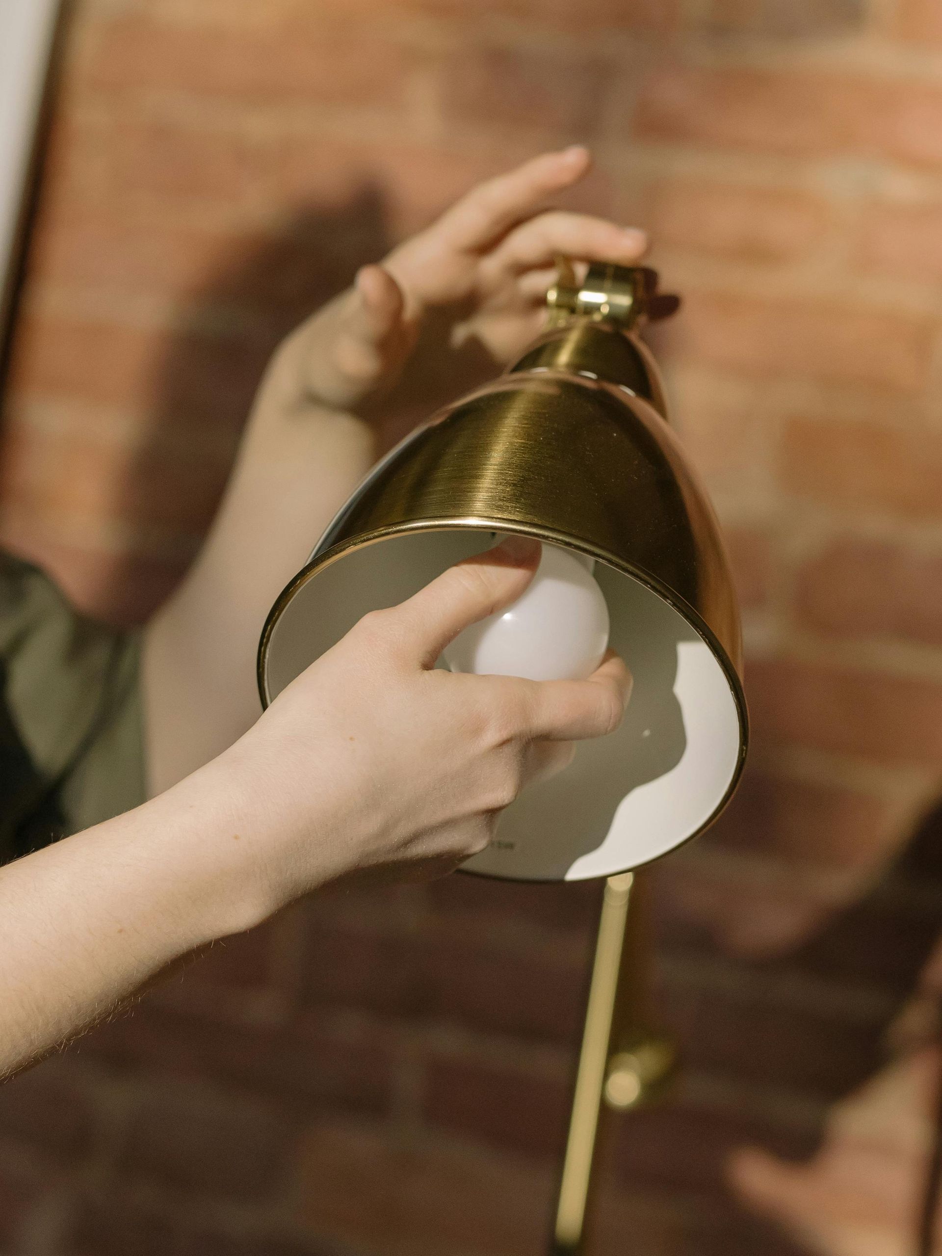 Hands reach into a metallic gold lamp shade to adjust a glowing, spherical white light bulb against a brick wall background.