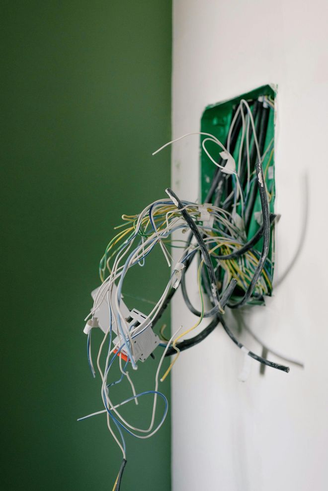 An open wall junction box with a messy bundle of exposed electrical wires hanging out against a green and white wall.