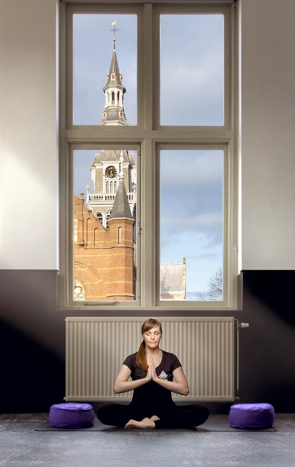 Een vrouw mediteert op een yogamat in een kamer met een groot raam en uitzicht op de stad.