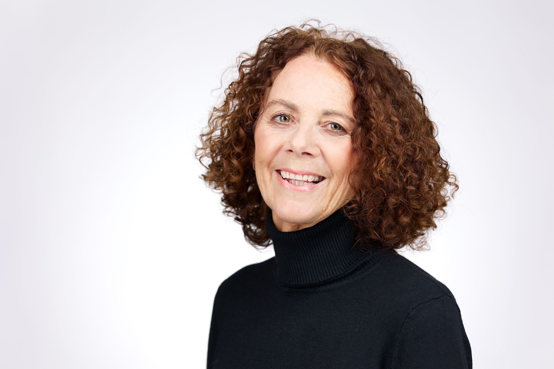 Lachende persoon met krullend haar in een zwarte coltrui tegen een witte achtergrond.