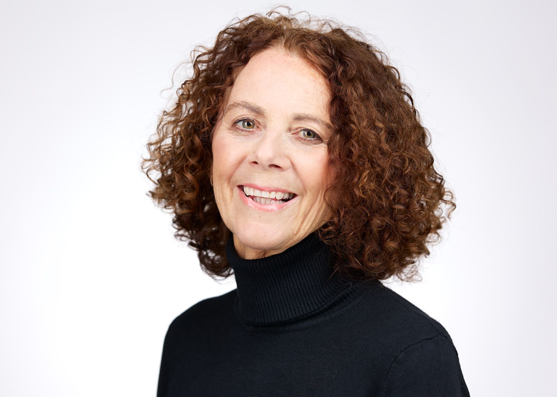 Een lachend persoon met krullend haar en een zwarte coltrui, afgebeeld tegen een witte achtergrond.