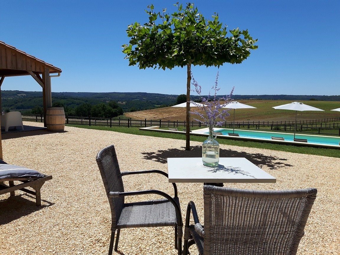 Buitenpatio met rieten stoelen, tafel, parasol en zwembad met uitzicht op een zonnige heuvel.