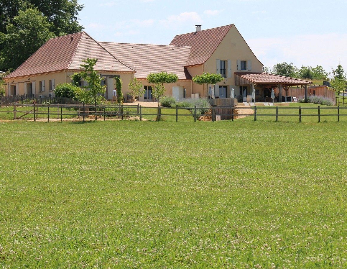 Groot beige huis met een rood dak naast een omheind groen veld op een zonnige dag.