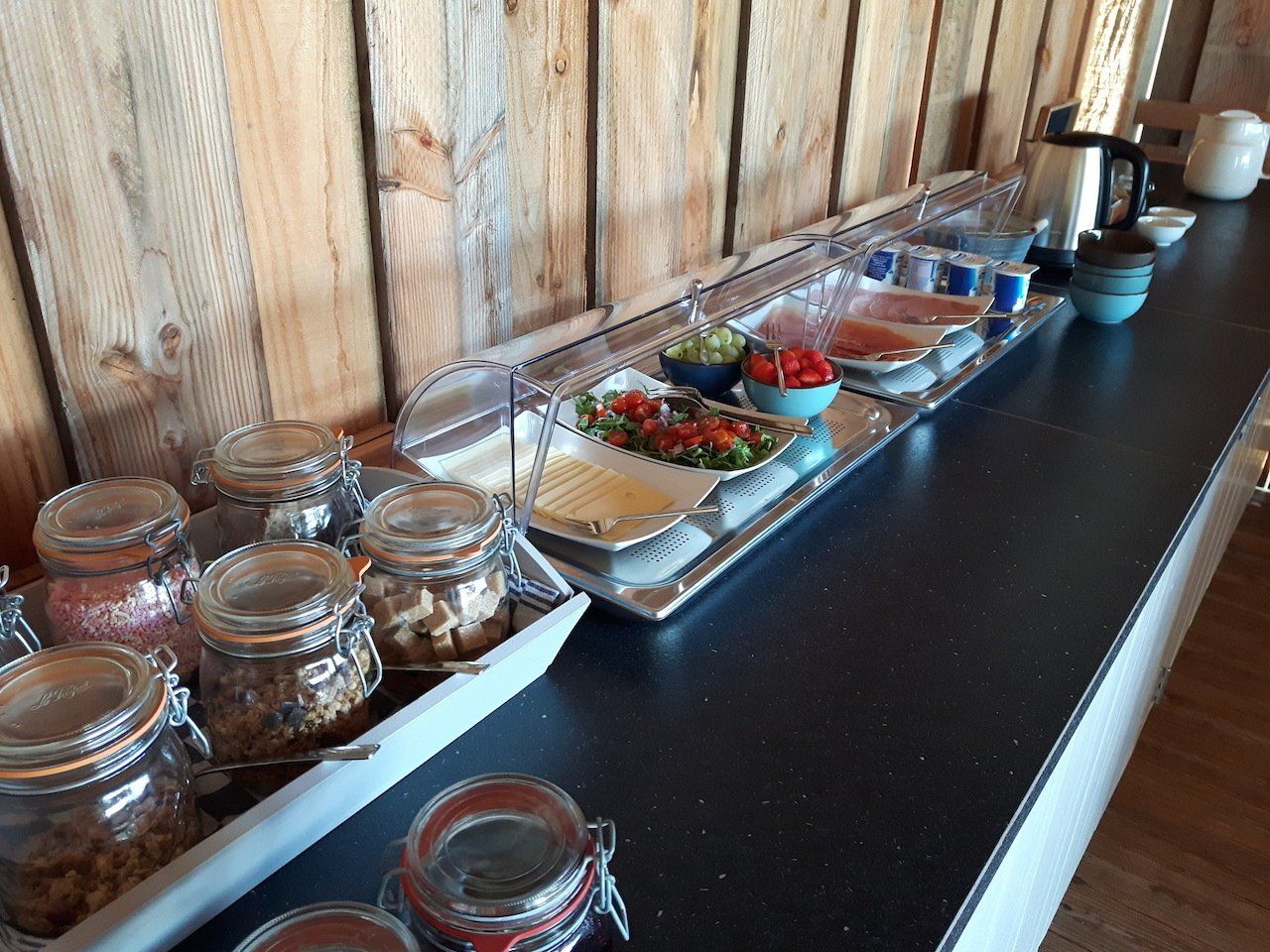 Buffettafel met potjes, salades en kruiden op een zwart aanrechtblad tegen een houten muur.