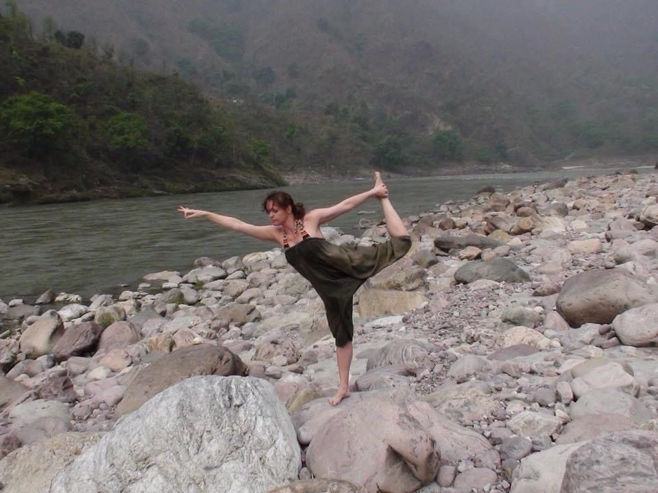 Een persoon die een yogahouding aanneemt op een rotsachtige rivieroever met mistige bergen op de achtergrond.