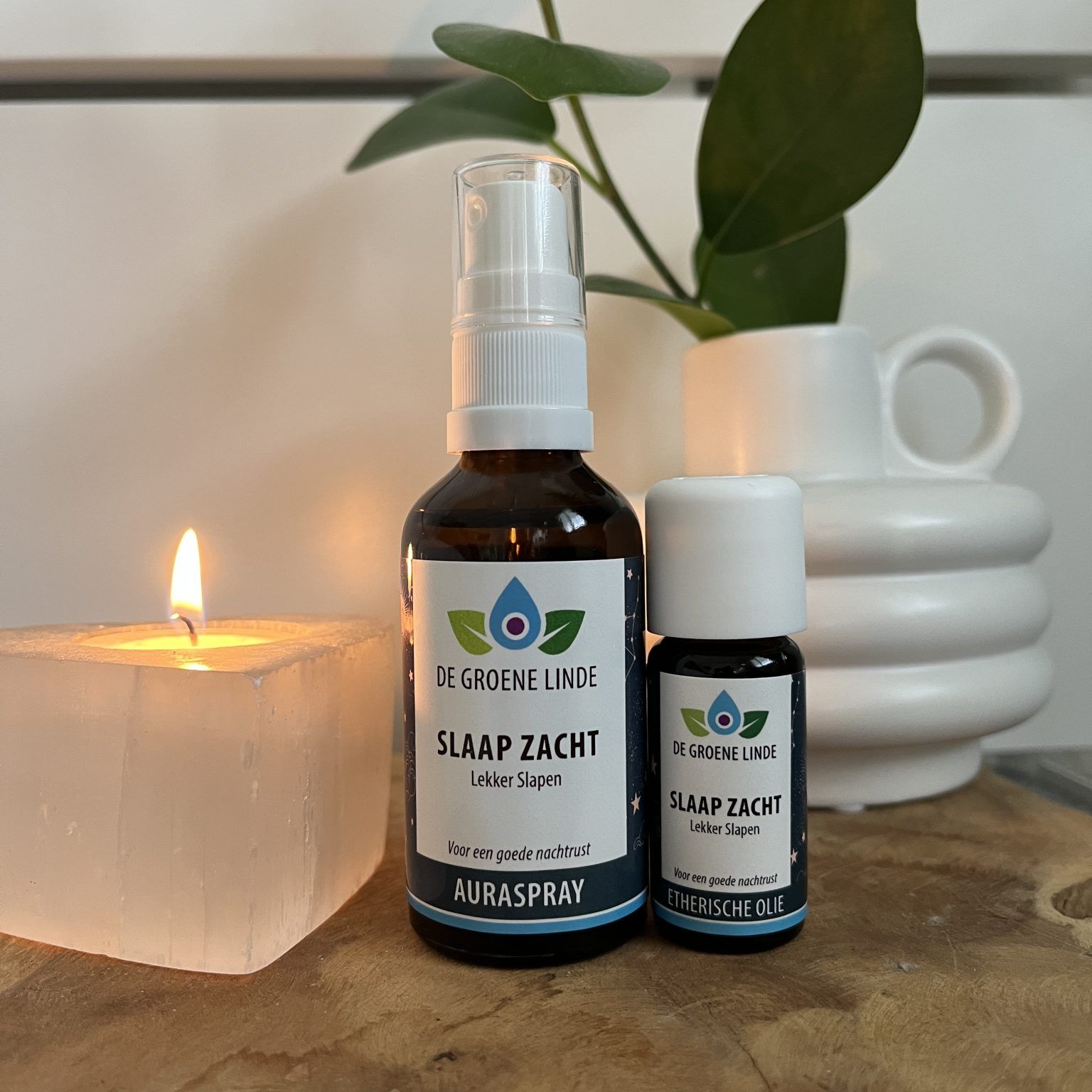 Een brandende kaars naast twee amberkleurige zeepflesjes op een tafel met een plant en opgestapelde witte kopjes.