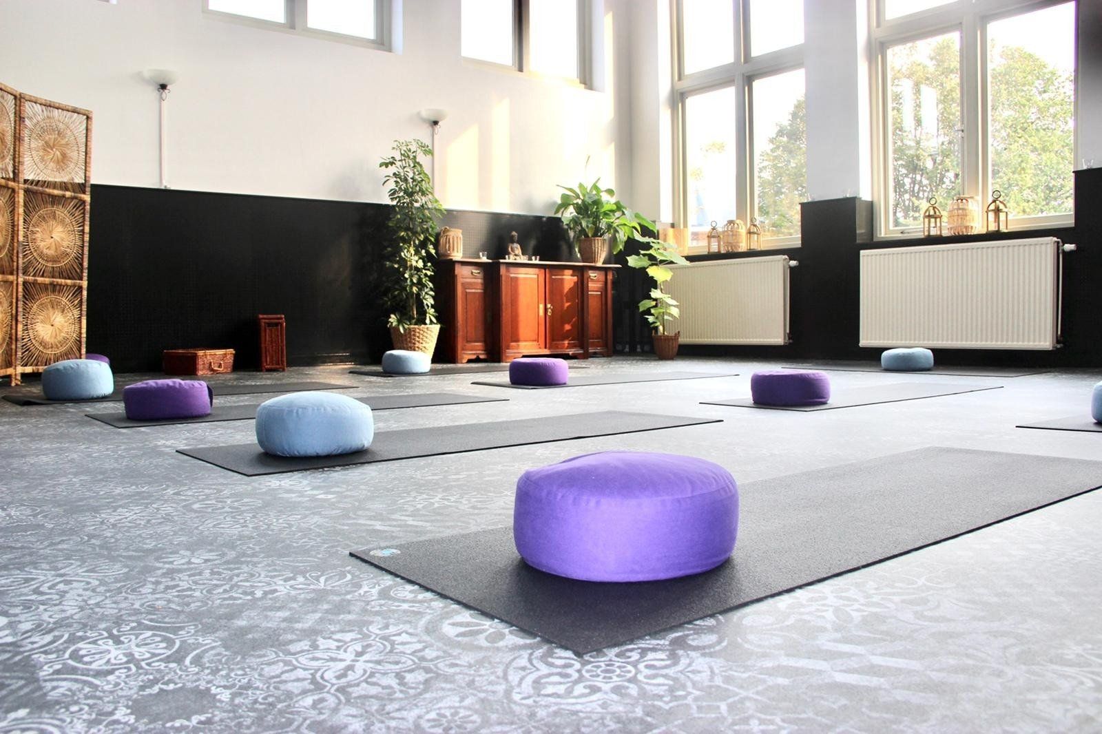Ruime yogastudio met matten en paarse kussens, badend in helder natuurlijk licht.