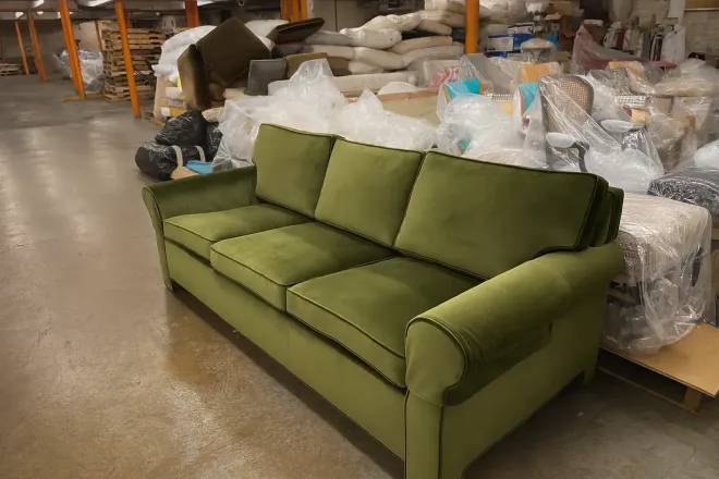 Green three-seat sofa in a warehouse aisle, surrounded by wrapped furniture and storage items.