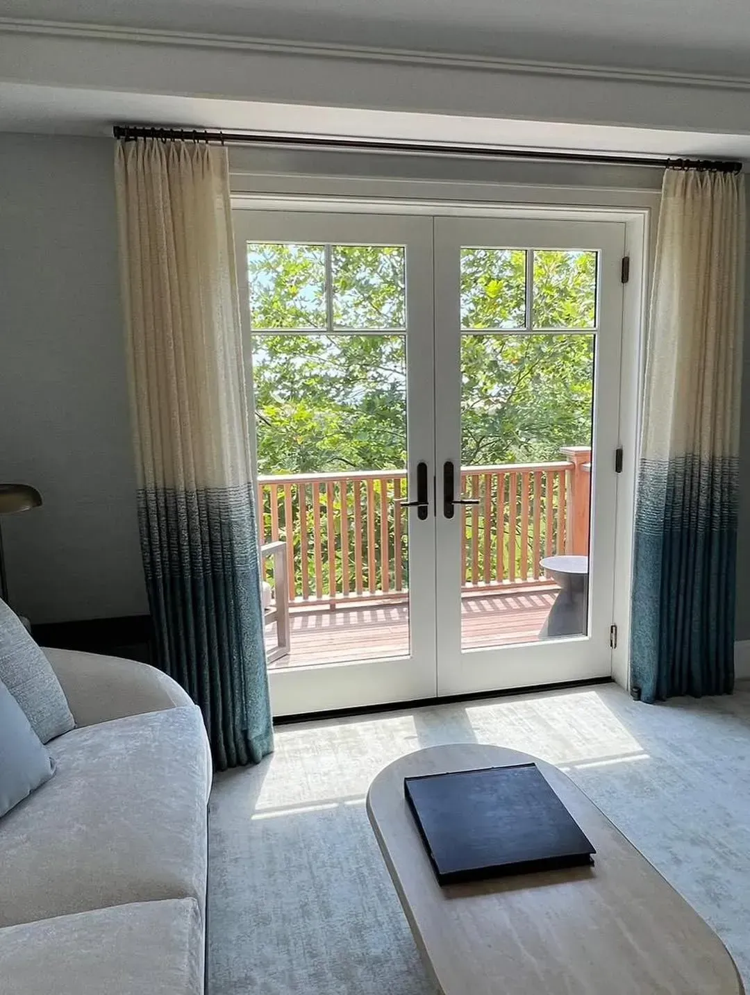 Bright room with open balcony doors, curtains, and a cushion on a light rug facing a wooded view