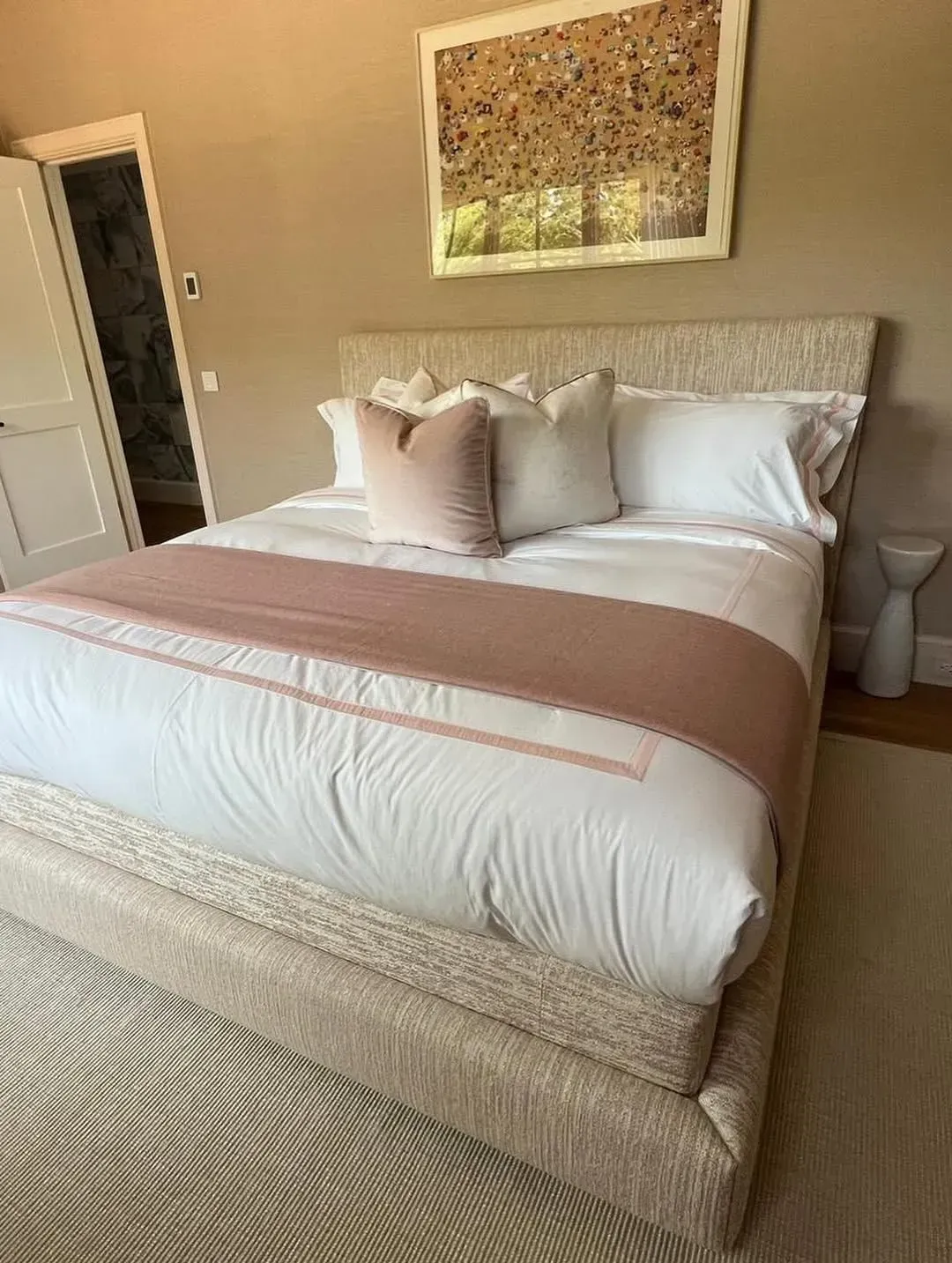 Neatly made bed with white bedding, blush throw, and framed art in a beige bedroom