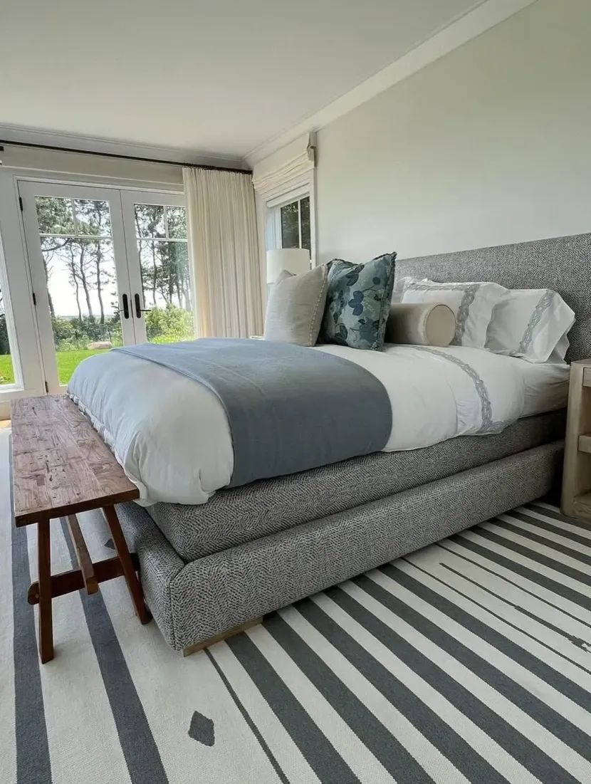 Bright bedroom with a large gray bed, striped rug, and glass doors opening to a green outdoor view.