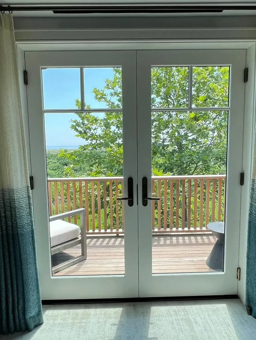 Sliding glass doors to a deck with wooden railing, green trees, and a bright blue sky beyond.