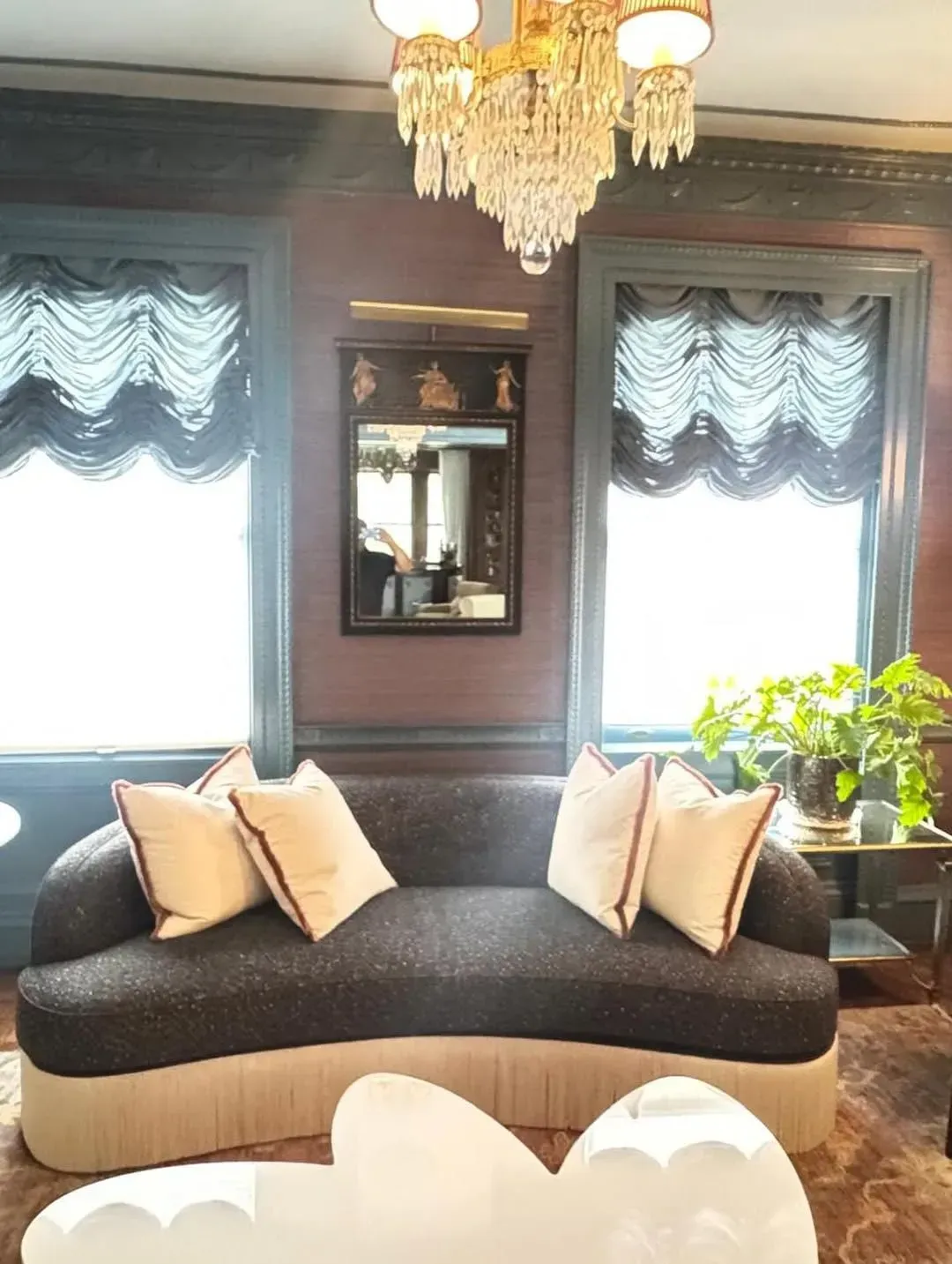 Elegant living room with a curved black sofa, cream pillows, windows with draped shades, chandelier, and a plant.