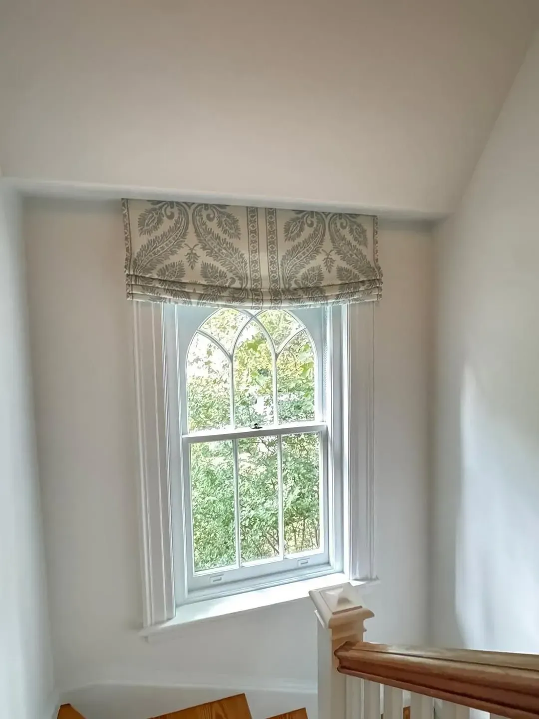 Stairwell window with patterned valance and white curtains above a wooden handrail