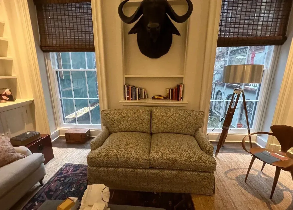 Cozy living room with a patterned loveseat, deer head wall decor, and large windows with bamboo shades.