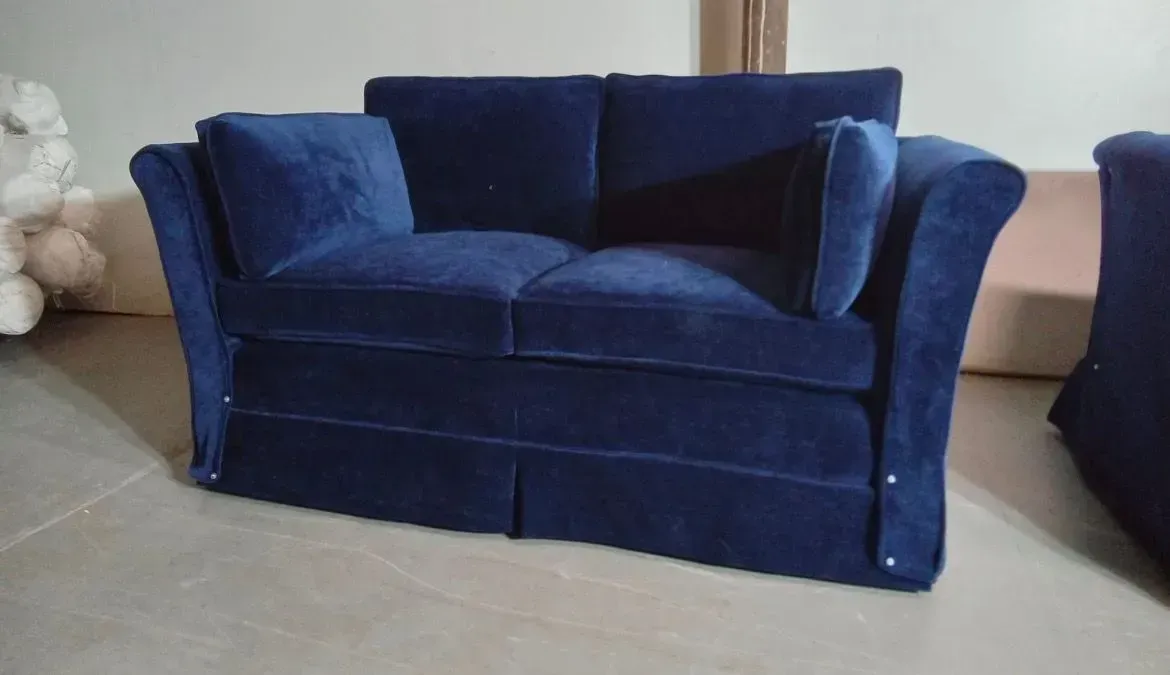 Blue velvet loveseat on a tiled floor in a living room, with a cream wall and another chair partially visible.