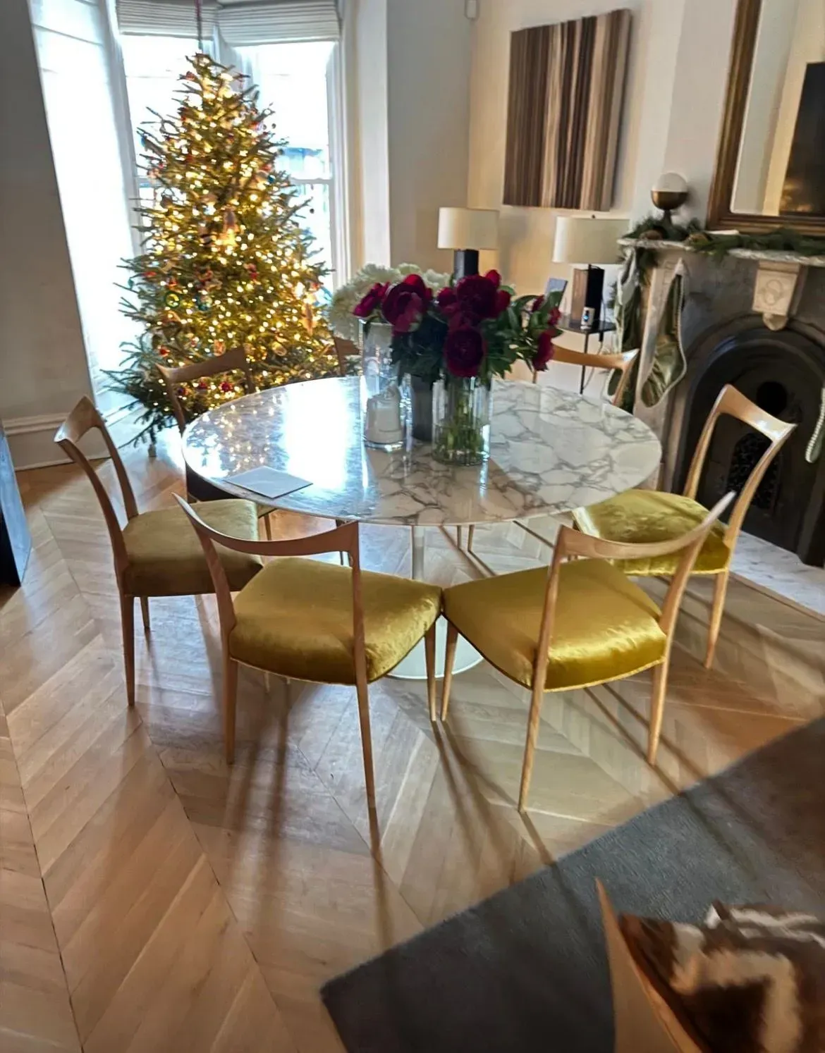 Dining room with lit Christmas tree, marble table, gold chairs, and flowers by a fireplace.