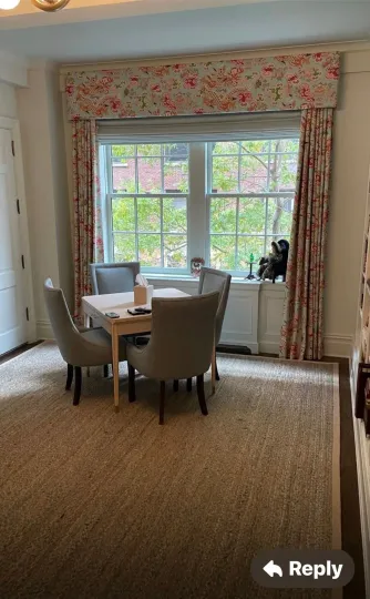 Dining area with round table, beige chairs, and floral curtains by a large window.