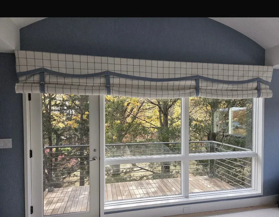 Window with blue valance and plaid blinds, looking out onto trees and a deck.