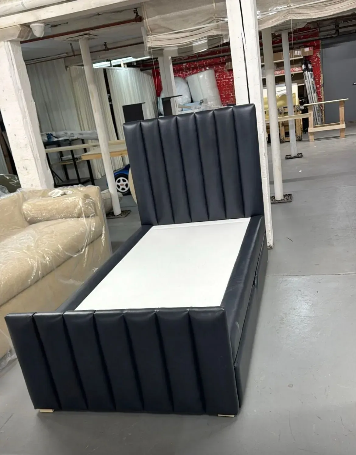 Black upholstered bed frame in a warehouse, with a white mattress base and tall vertical headboard.