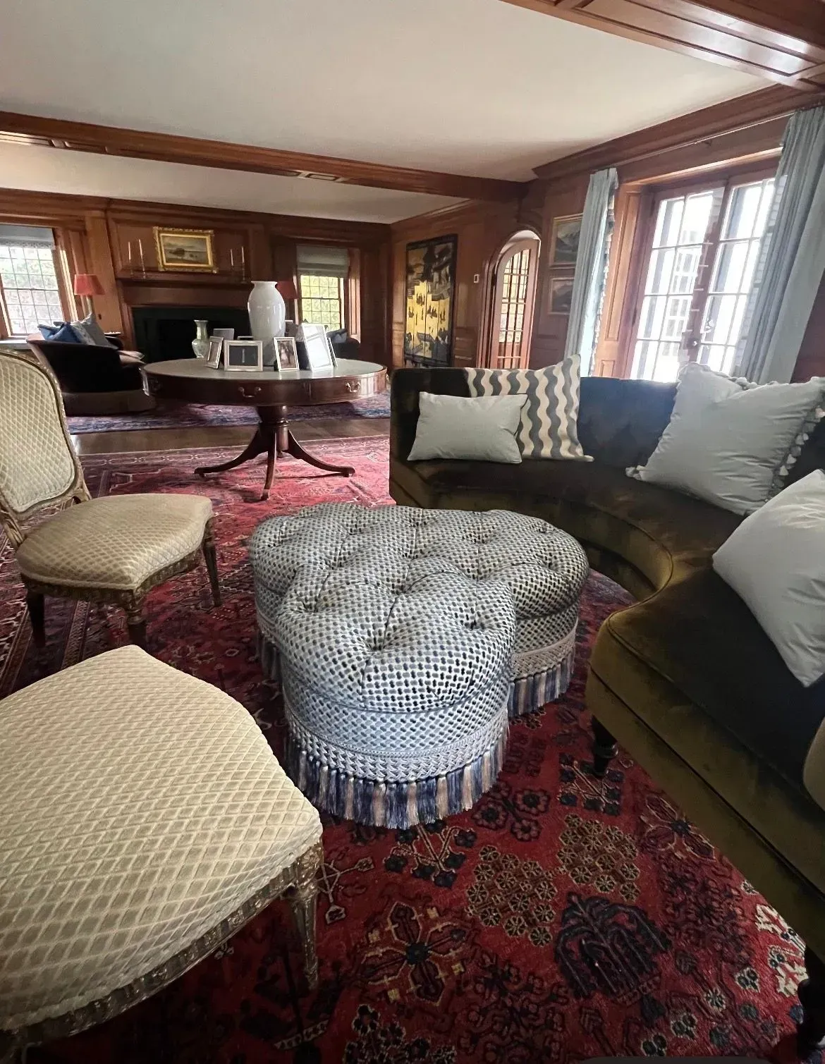 Cozy living room with dark wood paneling, red Persian rug, curved sofa, ottoman, and piano.