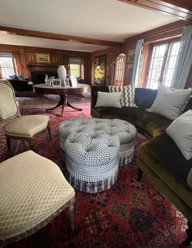 Cozy wood-paneled living room with upholstered chairs, sofas, ottomans, and a red patterned rug