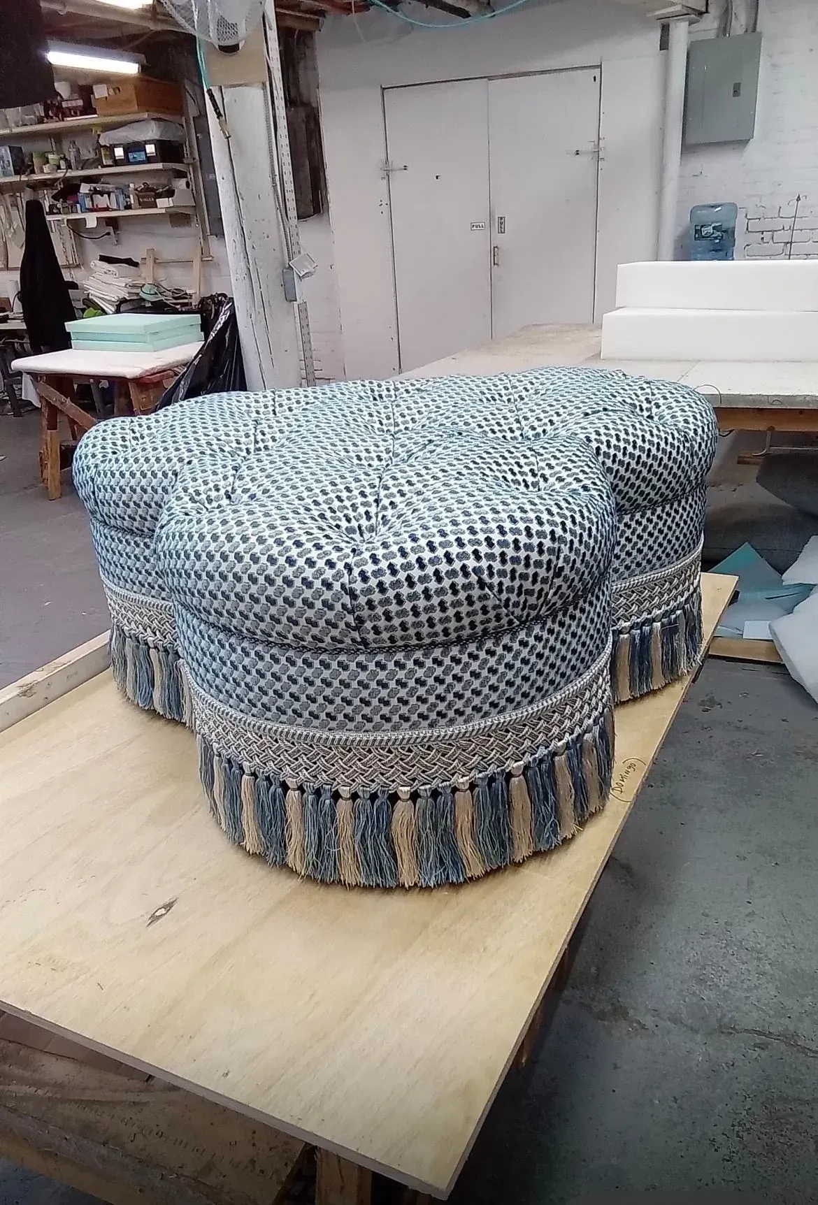 Blue patterned upholstered ottoman on a wooden table in a workshop, with a white sofa in the background
