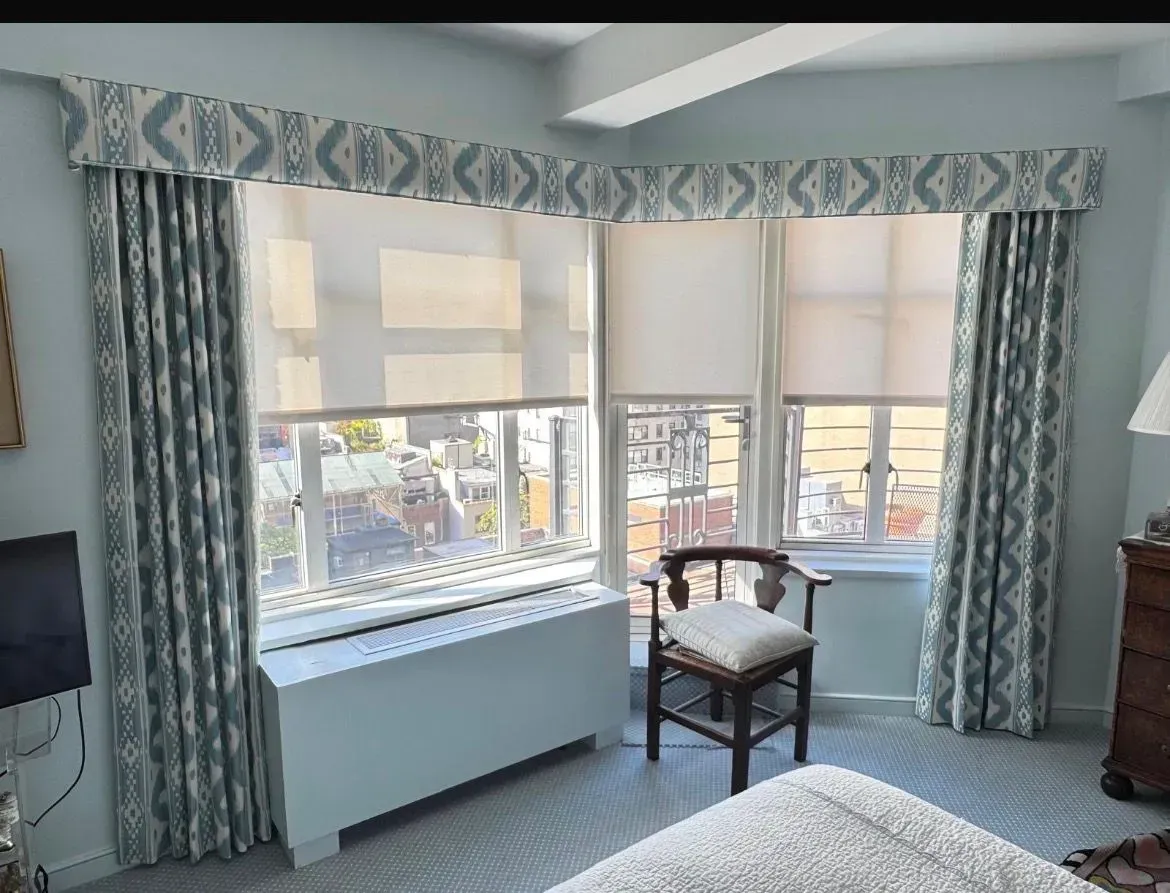 Bright bedroom with large bay windows, patterned drapes, and a chair by the window.
