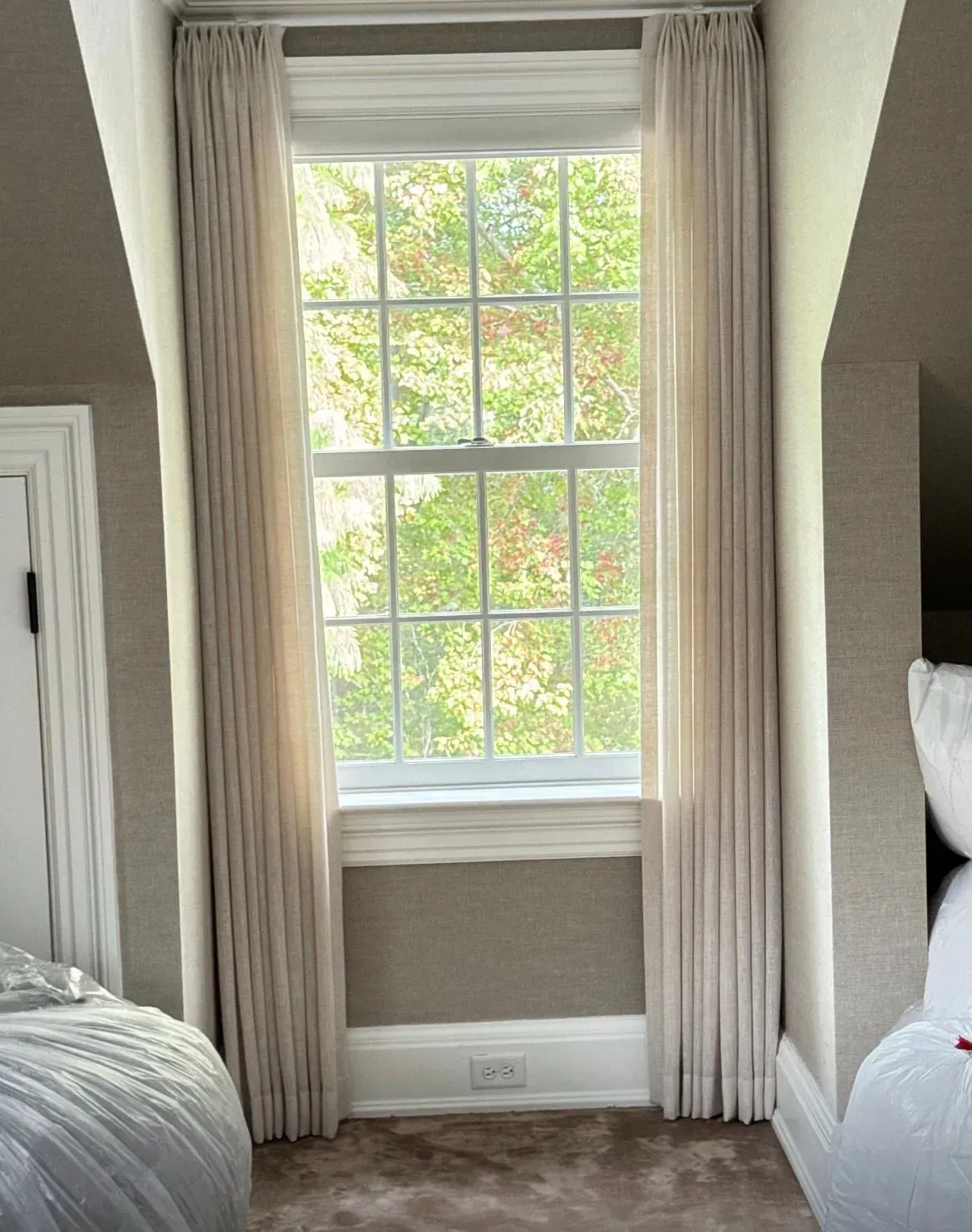 Bedroom window with beige curtains and a view of green trees outside