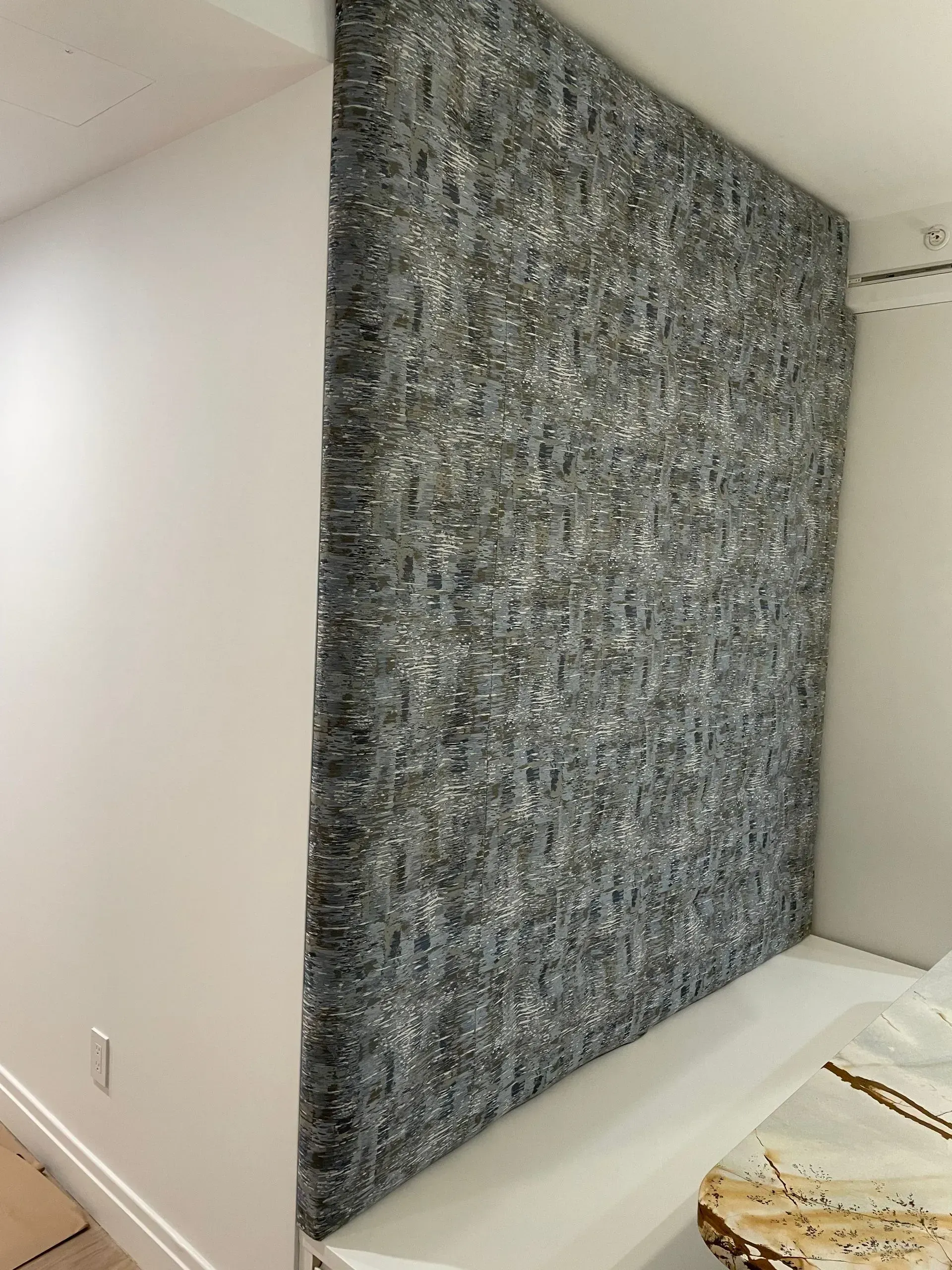 Large textured gray wall hanging in a minimalist white room beside a bed