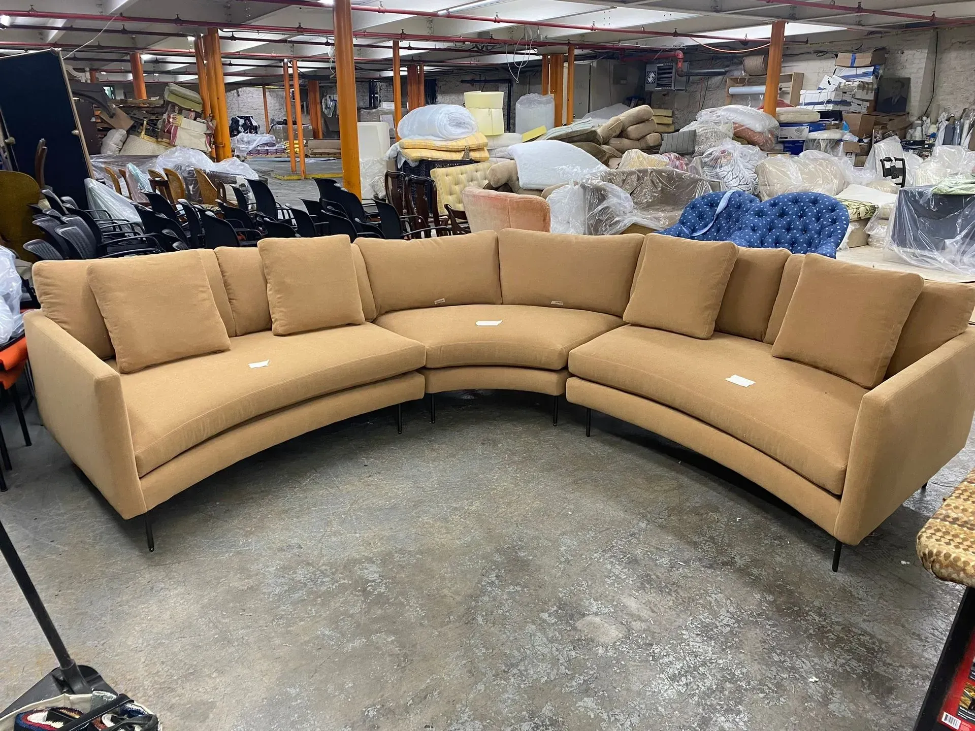 Curved beige sectional sofa in a warehouse showroom, surrounded by stacked furniture and supplies.