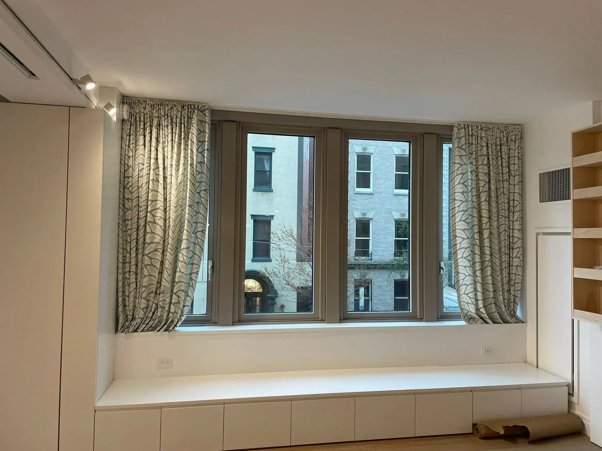 White bay window seat with built-in drawers and patterned curtains overlooking neighboring buildings