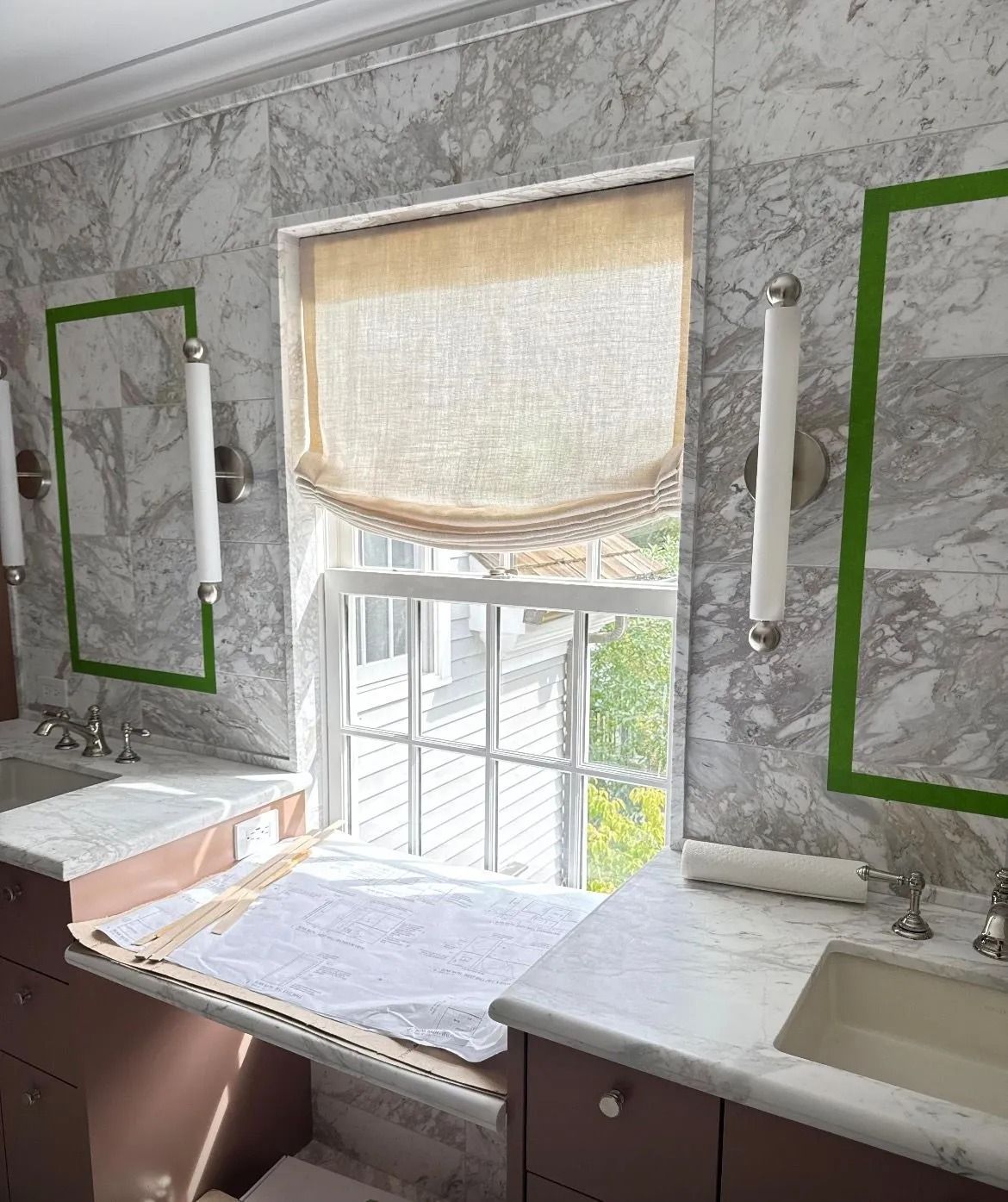 Bathroom vanity with marble-pattern wall, window, mirror lights, and sink beneath a roll-up shade