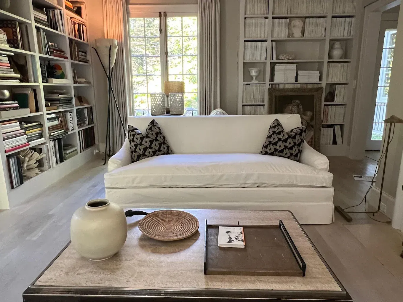 Cozy living room with white sofa, bookshelves, large windows, and a low coffee table with decor