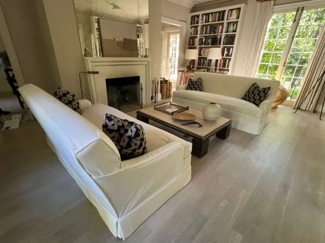 Bright living room with white sofas, fireplace, coffee table, bookshelves, and large windows