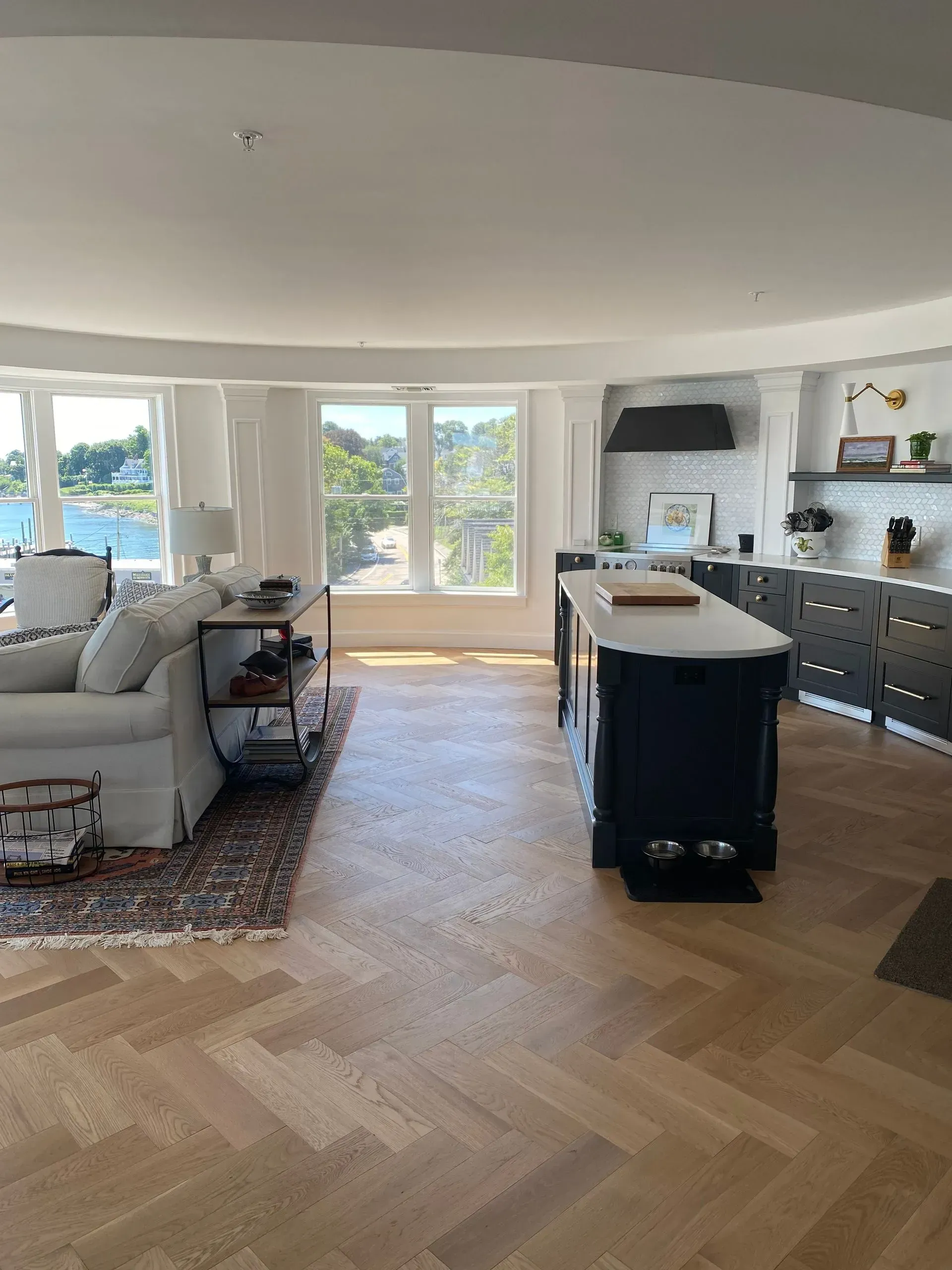 Bright open-plan living room and kitchen with herringbone wood floors, large windows, and a dark island counter