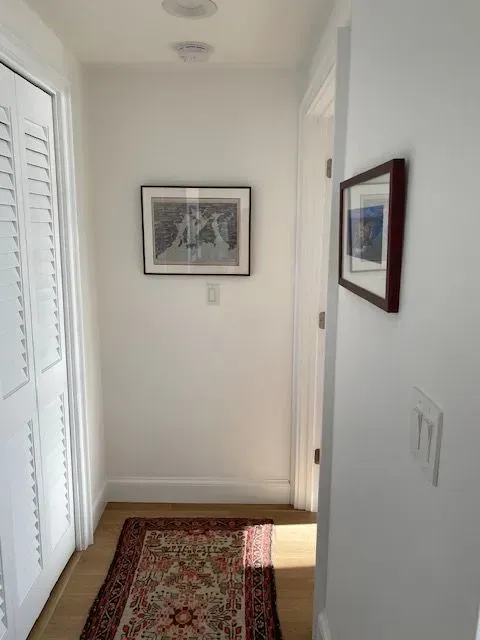 Narrow hallway with white walls, framed art, a patterned rug, and sunlight on the floor